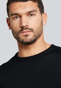 Schwarzer Strickpullover mit runder Ausschnitt und gerippten Abschlüssen, kombiniert mit einer Nahaufnahme der Gesichtszüge und des Bartes eines männlichen Models.