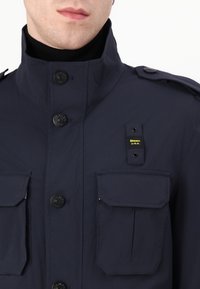 Donkerblauwe jas met een hoge kraag, knoopsluiting aan de voorkant, twee grote klepzakken en een merklabel op de borst. Gestructureerde stof met een matte afwerking.