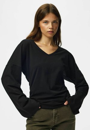 Jeune femme aux longs cheveux bruns portant un haut noir ample à col en V et manches longues, ainsi qu’un pantalon vert, posant avec les mains dans les poches.