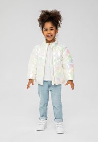 Chaqueta acolchada brillante e iridiscente con un collar alto, con un cierre frontal. Combinada con una camiseta blanca, jeans de color azul claro y zapatillas.