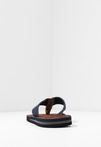 Sandal cu flip-flop navy, cu o curea împletită maro, partea superioară texturată, umplutură maro și o talpă din cauciuc negru cu accente gri.