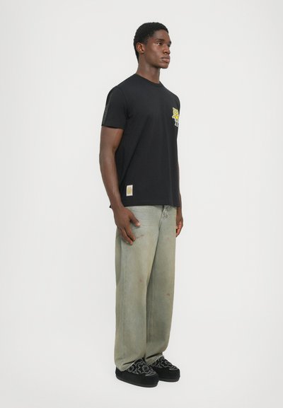 Black T-shirt featuring a yellow graphic design on the chest, paired with oversized light gray jeans and black patterned shoes.