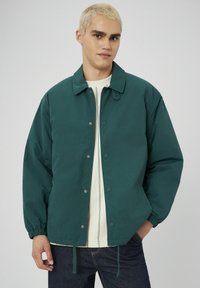 Veste légère teal avec des boutons-pression, des poignets élastiques et des cordons de serrage au bas. Portée sur un haut crème à rayures verticales.