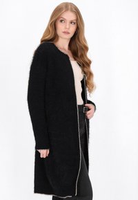 Schwarzer langer Cardigan aus strukturiertem, flauschigem Material. Mit offenem Frontbereich, kontrastierendem hellen Besatz und langen Ärmeln.