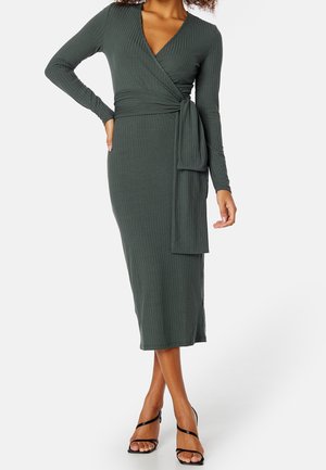 Femme portant une robe portefeuille côtelée à manches longues, vert foncé, avec un lien latéral, associée à des sandales noires à talons hauts et à lanières.