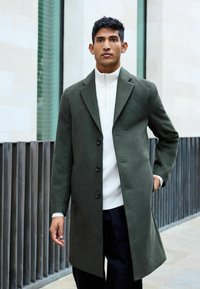 Grüner Wollmantel mit Reverskragen, einreihiger Knopfverschluss und seitlichen Taschen. Getragen über einem weißen strukturierten Stricktop.