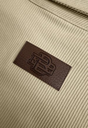 Tan stof met een gestructureerde weving heeft een donkerbruin leren label waar de initialen "PB" in zijn gedrukt. Stiksel omlijnt het label.