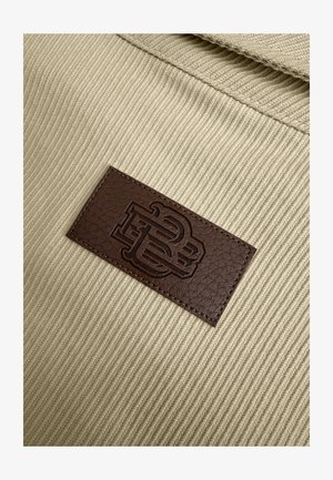 Tan stof met een gestructureerde weving heeft een donkerbruin leren label waar de initialen "PB" in zijn gedrukt. Stiksel omlijnt het label.