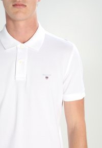 Polo blanc en tissu texturé, avec une patte de boutonnage à trois boutons et un logo brodé discret sur la poitrine gauche. Manches courtes.