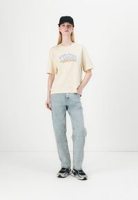 Crème katoenen t-shirt met blauwe en oranje tekst "TIMBS Authentic." Lichtblauwe spijkerbroek en zwarte pet maken de look compleet.