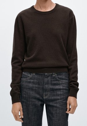 Personne portant un pull ajusté à manches longues marron foncé et un jean bleu foncé, debout devant un fond clair uni.