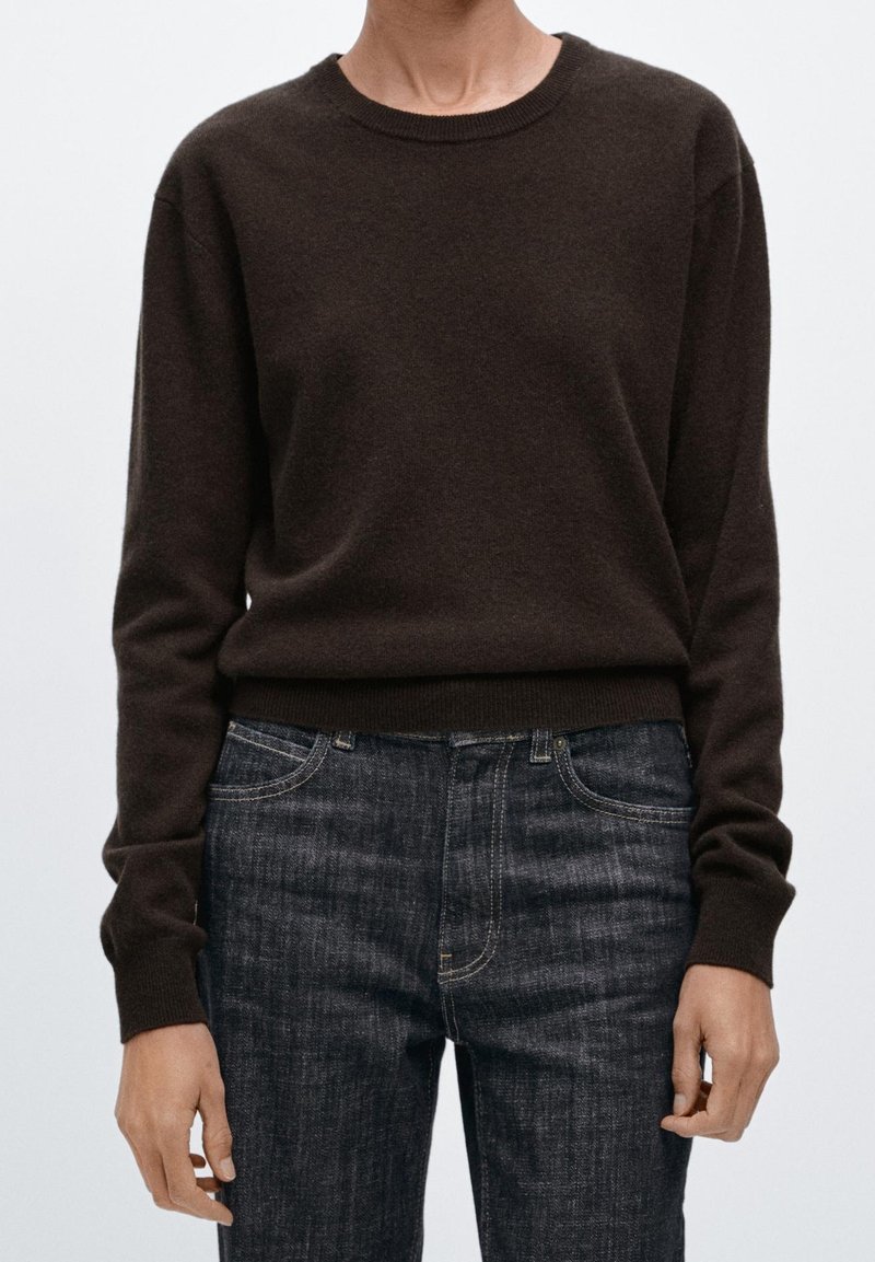 Personne portant un pull ajusté à manches longues marron foncé et un jean bleu foncé, debout devant un fond clair uni.
