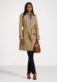 Beige trench coat with double-breasted buttons, waist tie, and epaulets. Paired with a blue and white striped shirt and dark jeans. Brown handbag.