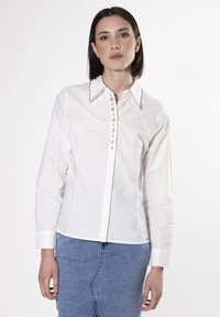 Camicia bianca con bottoni, maniche lunghe, colletto impreziosito e bottoni decorativi lungo il davanti. Tessuto liscio con una vestibilità sartoriale.