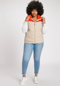 Gilet puffer beige con colletto in fleece arancione, chiusura a zip e due tasche frontali, indossato sopra una maglietta bianca a maniche lunghe e jeans blu.