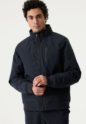 Hombre con cabello oscuro y rizado, vestido con una chaqueta negra con cremallera y bolsillos con cremallera en el pecho y las mangas, de pie frente a un fondo claro y sencillo.