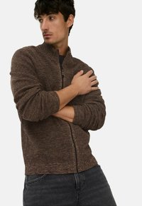 Brauner Reißverschluss-Pullover mit strukturiertem Strickmuster, hohem Kragen und gerippten Bündchen. Tragen über einem schwarzen Shirt mit dunklen Jeans.