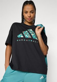 Zwarte oversized t-shirt met turquoise en grijs luipaardprintlogo; gecombineerd met turquoise shorts met witte Adidas-branding.