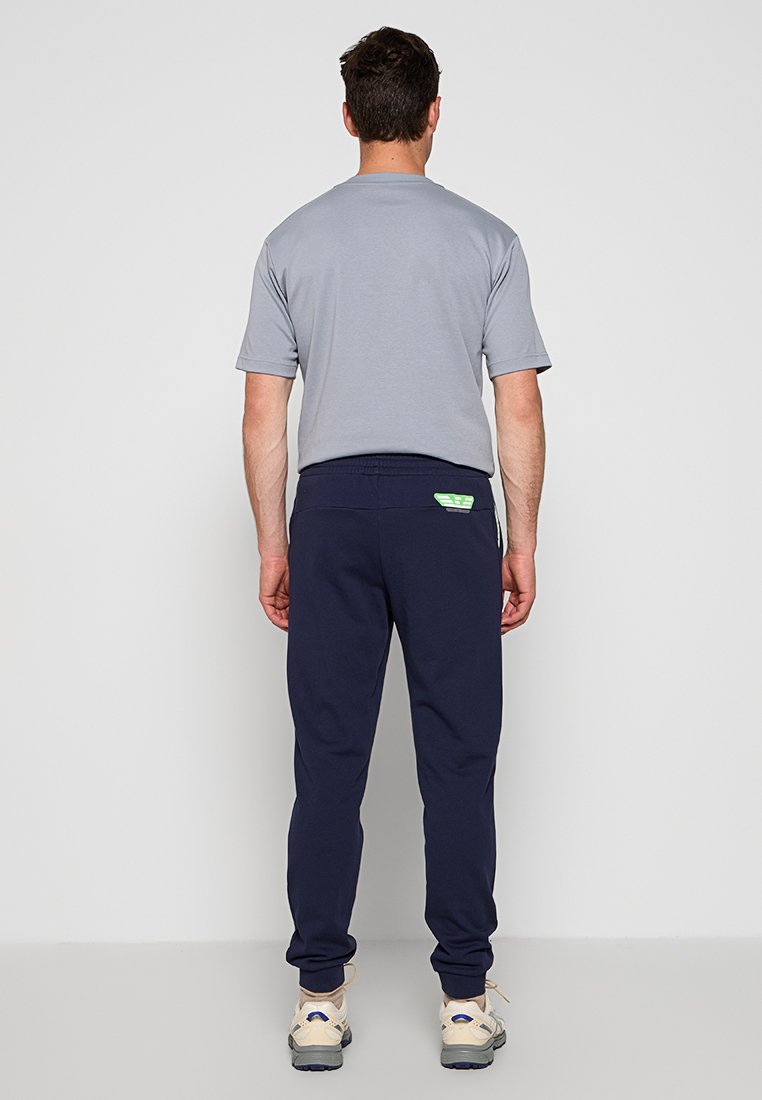 Szara koszulka z krótkim rękawem w połączeniu z granatowymi dresami; spodnie mają małą zieloną metkę; noszone z beżowymi butami sportowymi.