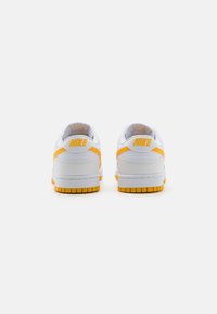 Weiße Nike-Sneaker mit gelben Akzenten. Sie verfügen über ein Obermaterial aus glattem Leder, einen gepolsterten Kragen und eine strukturierte Gummisohle. Branding an der Ferse.