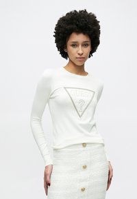 Λευκό μακρυμάνικο πουκάμισο με υφασμένο τριγωνικό λογότυπο «GUESS». Συνδυασμένο με λευκή φούστα που φέρει οριζόντια σχέδια και χρυσά κουμπιά.