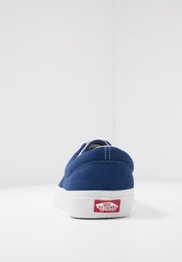 Blaue Wildleder-Slipper mit einer weißen Gummisohle und rotem Branding an der Ferse. Enthält Steppdetails und ein niedriges Profil-Design.