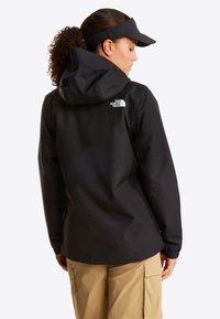 Γυναίκα με σγουρά μαλλιά σε κότσο, φορούσε μαύρο καπέλο με κουκούλα The North Face, μαύρη βίζιρα και μπεζ παντελόνι cargo, κοιτάζοντας προς τα πίσω.