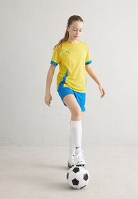 Fato de desporto da Puma amarelo e azul com uma camisa de manga curta, respirável, e calções a condizer. Jogador usa meias brancas e uma bola de futebol preta e branca.