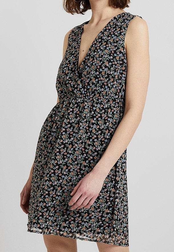 Femme portant une robe noire fleurie sans manches avec un col en V et une taille cintrée, debout devant un fond uni gris clair.