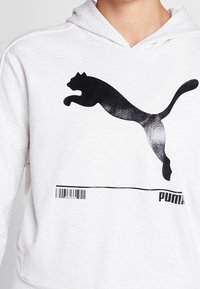 Persona che indossa una felpa bianca con un grande logo di un gatto nero che salta e un design a codice a barre sopra la parola "PUMA" sul davanti.