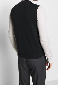 Person wearing a black sleeveless sweater over a light beige long-sleeve top and dark gray pants, shown from the back.