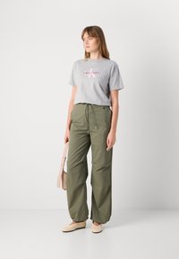 Camiseta de algodón gris con logo rosa de "Calvin Klein", combinada con pantalones holgados verde oliva y zapatos planos de color beige claro. La textura es suave.