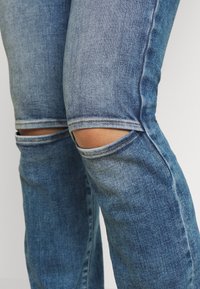 Närbild av slitna blå denimjeans med horisontella rivningar vid knäna som visar huden under mot en enkel bakgrund.