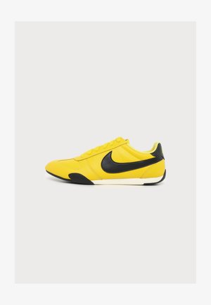 Žlutá teniska s černým logem Nike, černými detaily na špičce a patě, bílá střední podrážka, pohled z boku na jednoduchém pozadí.