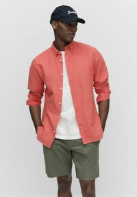 Koraalrood overhemd met opgerolde mouwen over een wit T-shirt, gecombineerd met groene shorts en een donkerblauwe baseballcap.