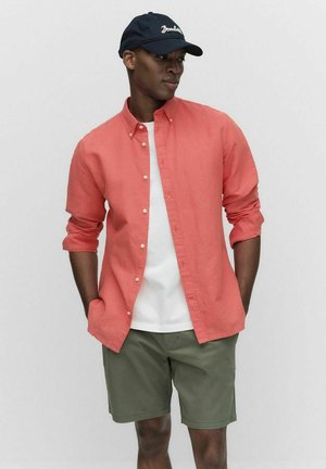 Koraalrood overhemd met opgerolde mouwen over een wit T-shirt, gecombineerd met groene shorts en een donkerblauwe baseballcap.