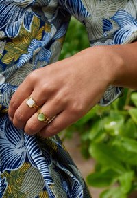 Zwei Goldringe an einer Hand, einer mit einem weißen rechteckigen Stein und der andere mit einem grünen runden Stein, vor einem gemusterten Stoffhintergrund.