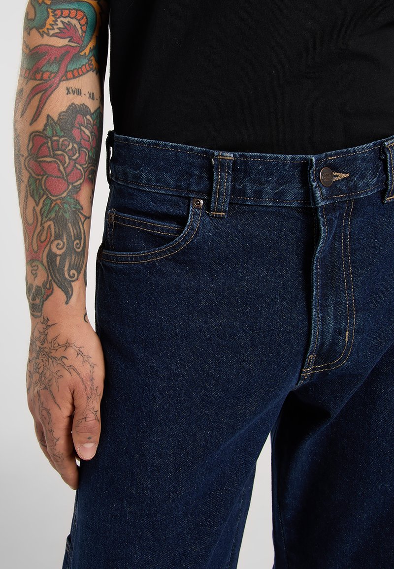 Tatueret venstre arm med farverige designs og sorte blækstreger hviler ved siden af mørkeblå denimjeans og en sort skjorte.