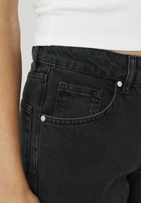 Jean en denim noir avec une finition texturée, présentant une poche avant, des rivets en métal et des coutures visibles le long de la taille.