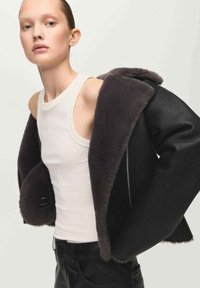 Czarna kurtka z czarnej sztucznej futra z miękką szarą podszewką, z dużym kołnierzem i zamkiem błyskawicznym, noszona na białym topie bez rękawów i czarnych spodniach.