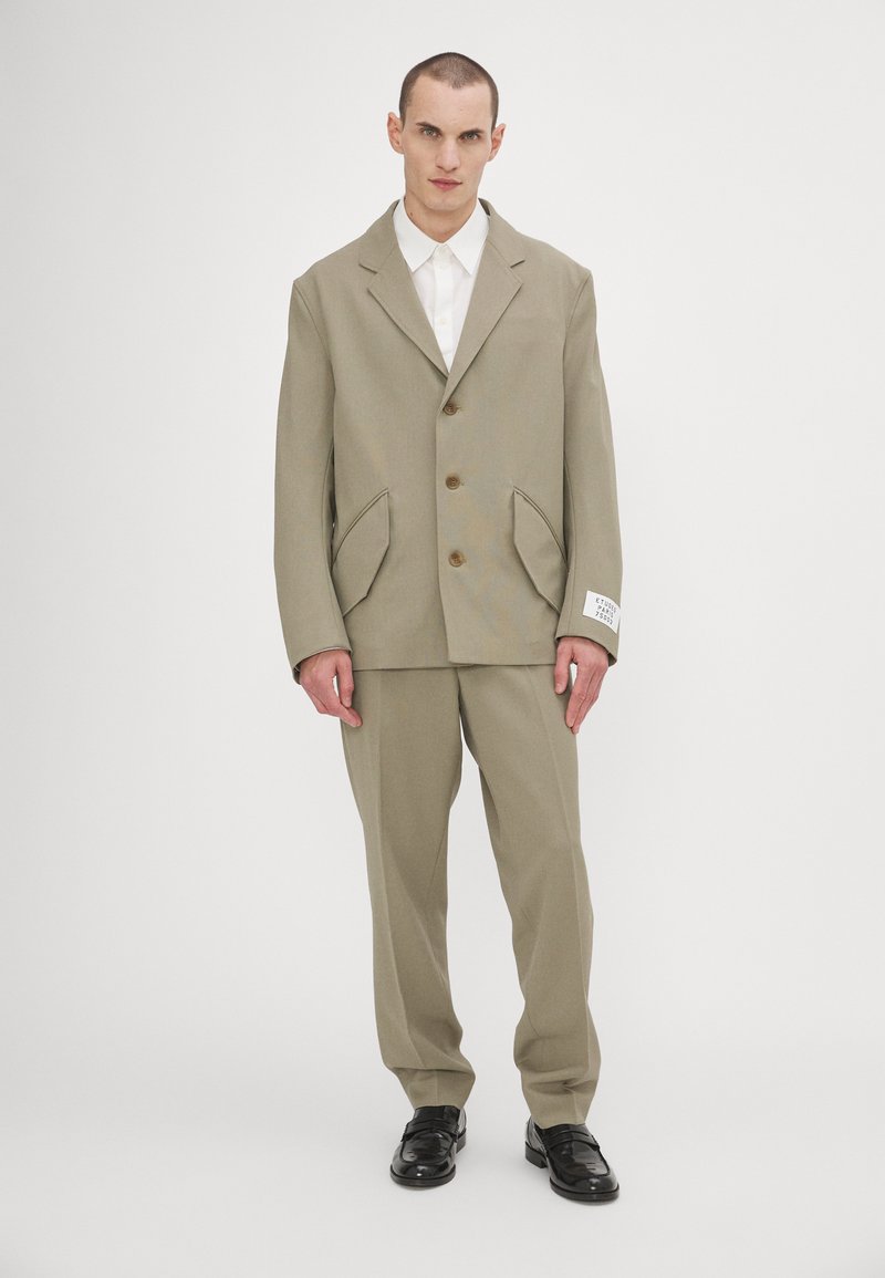 Beige maßgeschneiderter Anzug mit einem doppelreihigen Blazer und einzigartigen Taschen, kombiniert mit passenden Hosen und schwarzen Schuhen.