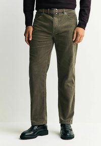 Olivgrüne Cordhosen mit geradem Bein, vorne mit Taschen, abgesteppten Nähten und einem braunen Ledergürtel. Schwarze Loafers werden getragen.