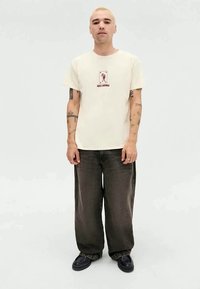 Beige bomulds t-shirt med korte ærmer, prydet med et rødt grafisk print. Sammen med mørke, løse denim jeans og sorte sko.