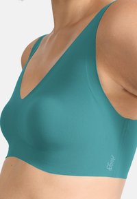 Teal seamless bra with a deep V-neck, wide shoulder straps, and a smooth texture. Features a small logo on the side.