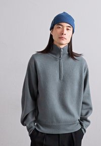Gray knit sweatshirt with a high collar and half-zipper, featuring long sleeves and a ribbed hem, paired with a blue beanie.
