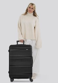 Mujer con un suéter de cuello alto beige y pantalones blancos, sosteniendo una maleta rígida negra con textura, de cuatro ruedas y con un mango extensible.