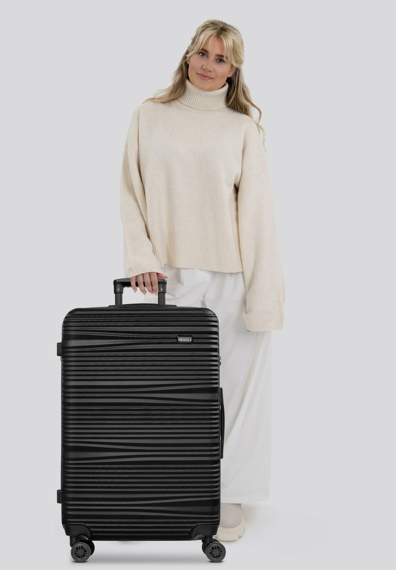 Mujer con un suéter de cuello alto beige y pantalones blancos, sosteniendo una maleta rígida negra con textura, de cuatro ruedas y con un mango extensible.