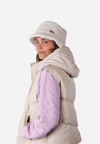Beige Steppweste mit Kapuze, pinkes strukturiertes Langarmoberteil und weißer flauschiger Bucket Hat mit einem kleinen braunen Etikett auf dem Hut.