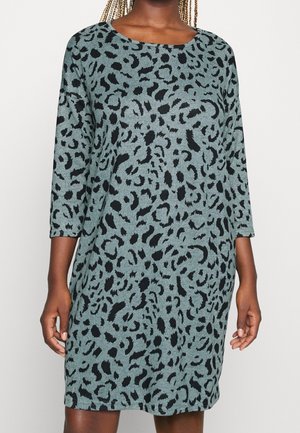 Femme portant une robe ample longueur genoux avec des manches trois-quarts et un motif animalier gris et noir.