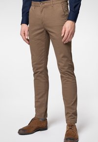 Homme portant un pantalon marron moulant et des chaussures à lacets en daim marron devant un fond uni.